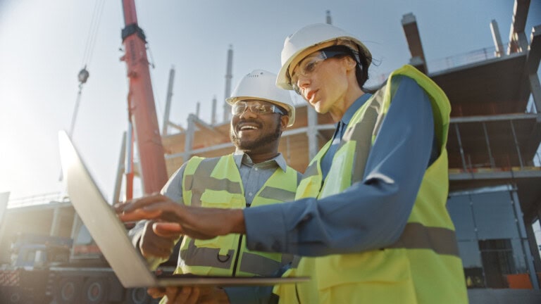 Two construction workers wearing safety gear review construction expenses on a laptop at an active construction site with a crane and building framework in the background.
