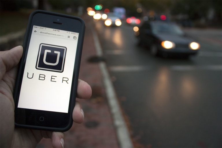 25958_11_06_uber_story_south58f-1200x800.jpeg A hand holding a smartphone displaying the Uber app in front of a blurred street with moving cars at dusk.