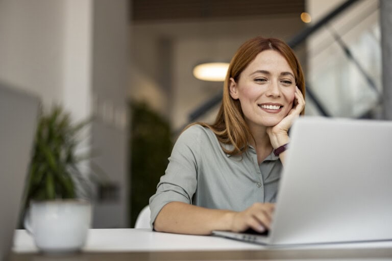 A woman with red hair sits at a desk, smiling while looking at a laptop. looking up Accounting software integrations that integrate seamlessly with ERP software
