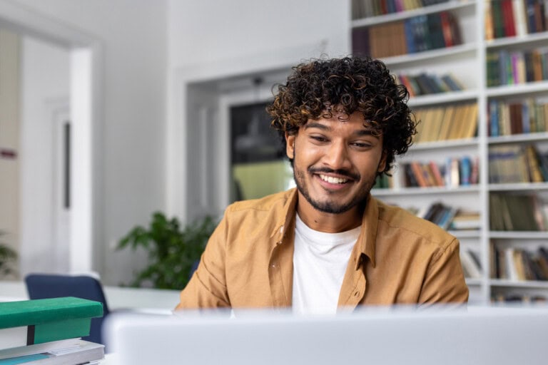 Young businessman works with accounting workflow automationing on laptop with a cheerful smile A person with curly hair is smiling while looking at a laptop, seated in a bright room with bookshelves in the background, likely exploring AI advancements in finance workflow automation.