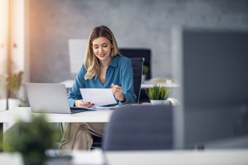 A woman in a blue shirt sits at a desk in an office, looking at a document and using a laptop.