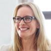 A woman with long blonde hair and glasses smiles while looking slightly to the side, with a window in the background.