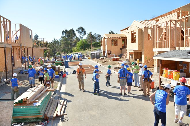 A group of people wearing blue shirts and hard hats work on multiple house constructions under a sunny sky.