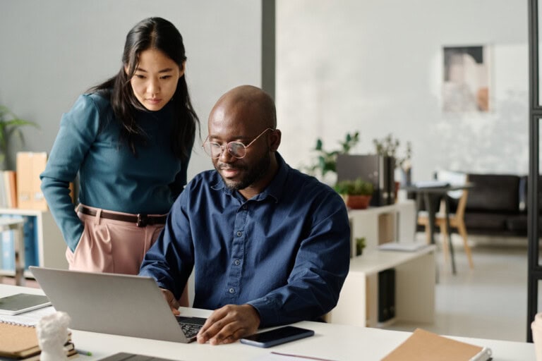 Asian Woman And Black Man discussing automated reconciliation A man sits at a desk working on a laptop while a woman stands beside him, looking at the screen in a modern office setting.