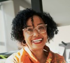 A woman with curly hair and glasses smiles, wearing an orange shirt, seated indoors with sunlight in the background.