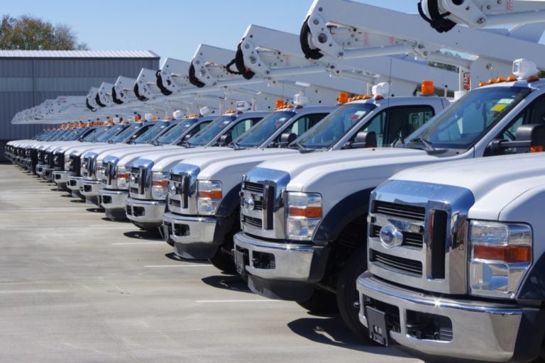 A row of white utility bucket trucks parked in a lot, with hydraulic lifts extended.