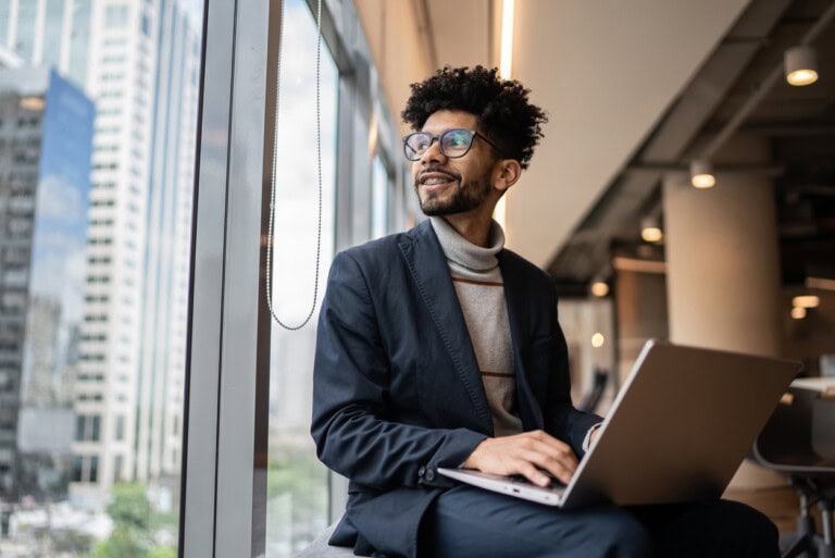 A man in business attire sits by a window in a modern office, using a laptop and choosing the right expense management application
