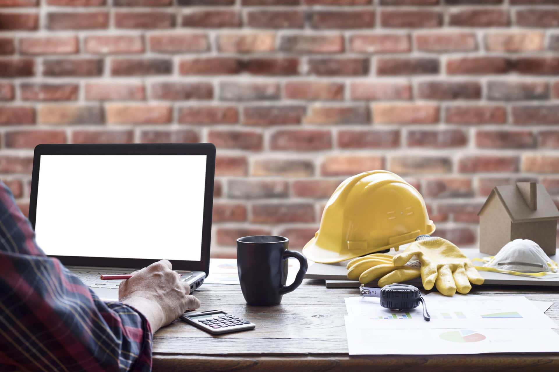 A desk with a laptop, coffee mug, calculator, yellow hard hat, gloves, measuring tape, and papers in front of a brick wall.