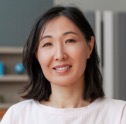 A woman with shoulder-length dark hair and a light-colored top smiles at the camera, with blurred shelves and objects in the background.
