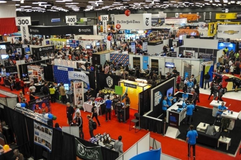 A busy convention hall with numerous booths, banners, and attendees interacting and exploring displays.