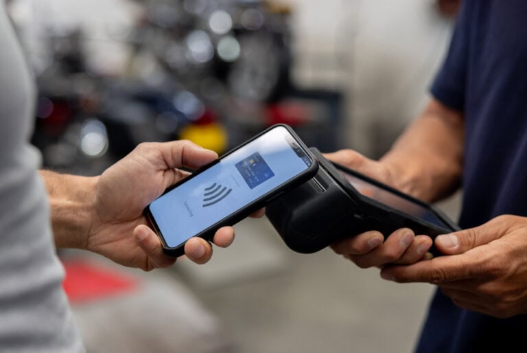 A person holds a smartphone near a card reader for contactless payment, while another person holds the terminal.