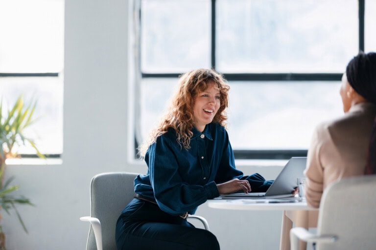 Two Women Collaborating in a Modern Office Setting A woman with long curly hair sits at a table with a laptop, smiling and talking about employee expense reimbursement.