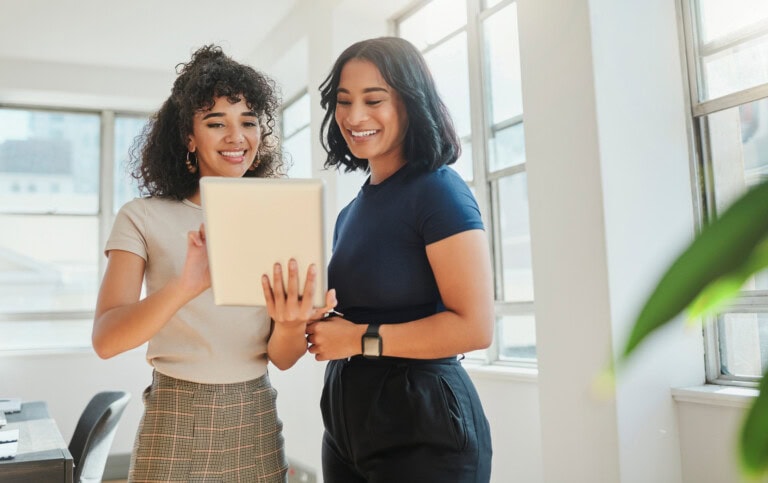 Two people stand in a well-lit room, smiling as they look at a tablet displaying expense reporting software 101. One is wearing a beige shirt and plaid skirt, while the other sports a dark shirt and pants.