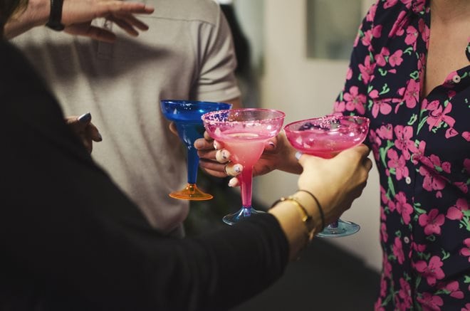 Three people holding colorful margarita glasses filled with pink drinks, toasting at a casual gathering.