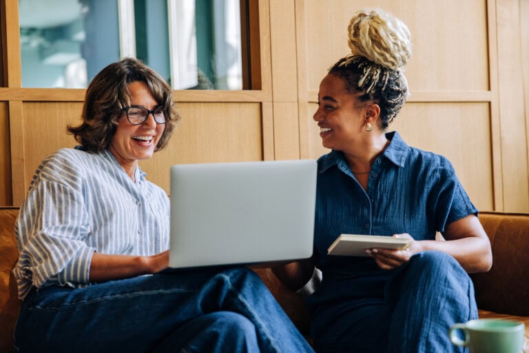 Two women smiling while using a laptop and notepad indoors Financial reporting for growing teams 4 steps to stay ahead