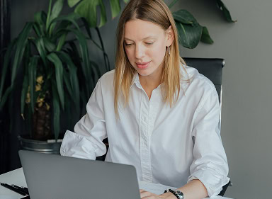 Frame 1171276253 Woman in a white shirt sitting at a desk and working on a laptop, with a plant in the background.