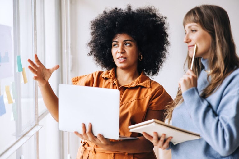 Female workers brainstorming about financial controlsin an office Two women stand by a window, collaborating; one gestures while holding a laptop displaying financial controls, the other holds a notebook and pen, both facing sticky notes on the glass.
