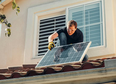 Group 1171275100 (1) A person wearing gloves and safety glasses installs a solar panel on the roof of a house with white window shutters.
