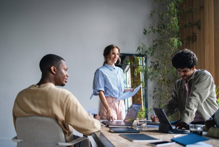 Three people at a modern office desk demonstrate back-office optimization, with one standing and holding papers while two others work on laptops.