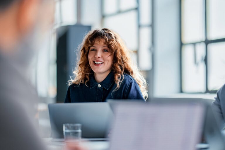 Woman with curly hair sits at a table in a bright office, smiling, with an open laptop in front of her, seamlessly managing tasks using expense management software as blurred colleagues are seen in the foreground.