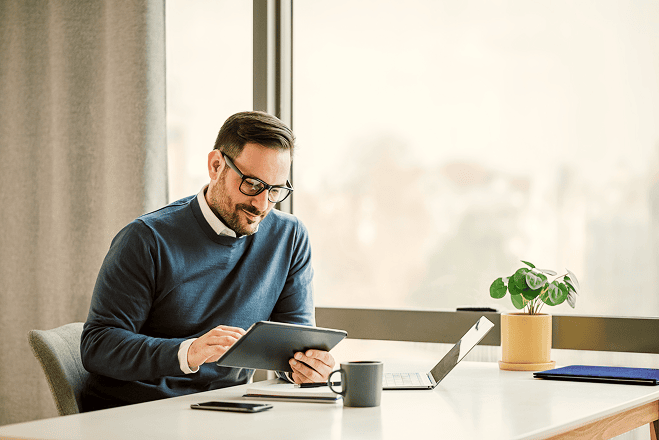 A man in glasses sits at a desk by a window using a tablet with spend management software. A laptop, smartphone, mug, and small potted plant are arranged neatly on the desk.