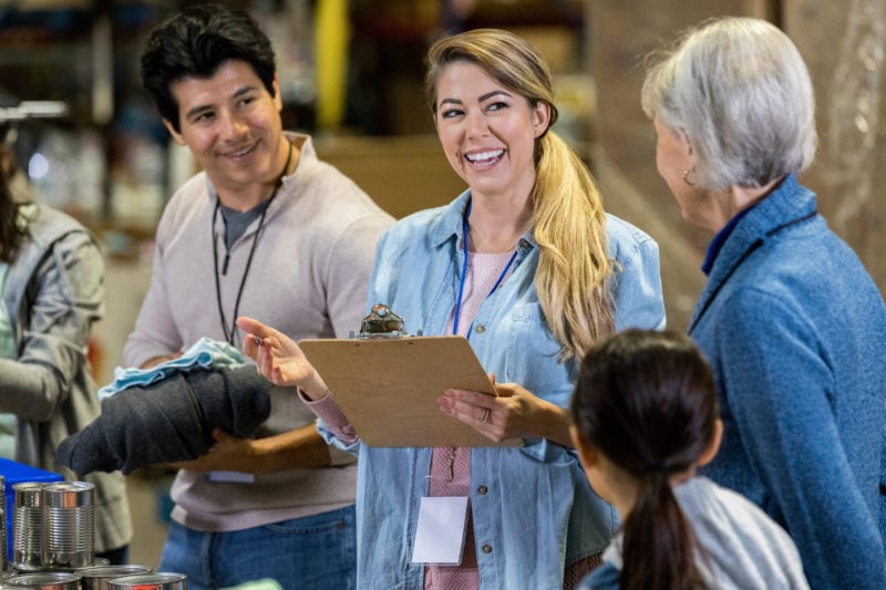 A smiling mid adult woman stands in a food bank warehouse and holds a clipboard.  She is discussing procedure with a couple of volunteers.