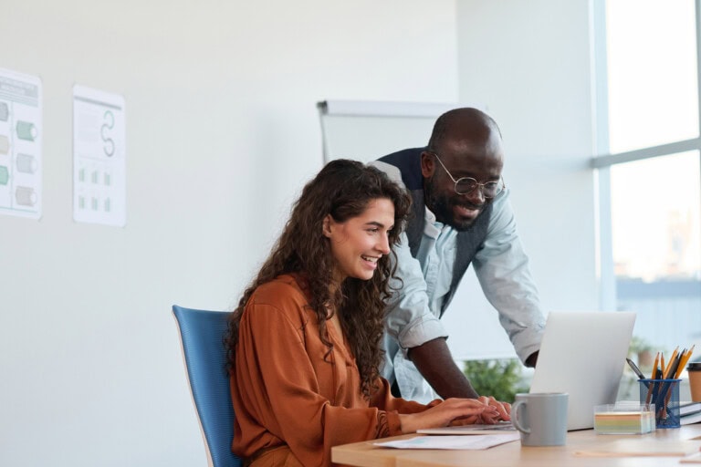 A woman sits at a desk working on a laptop while a man stands beside her, both looking at the screen and comparing PEX vs WEX