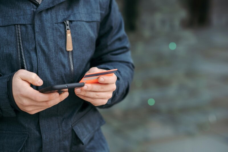 Person in a dark jacket holds a smartphone and a credit card, poised for online payment, while embracing the ease of automated reconciliation.