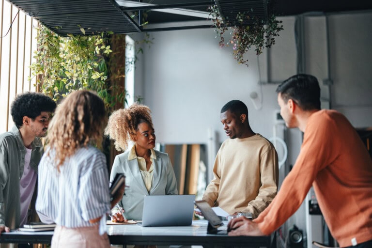 A Group Of Multiethnic Businesspeople Working Together In The Office A group of five people stands around a table with a laptop, engaged in discussion about streamlining back office operations through Ai and automation inside a room with hanging plants.