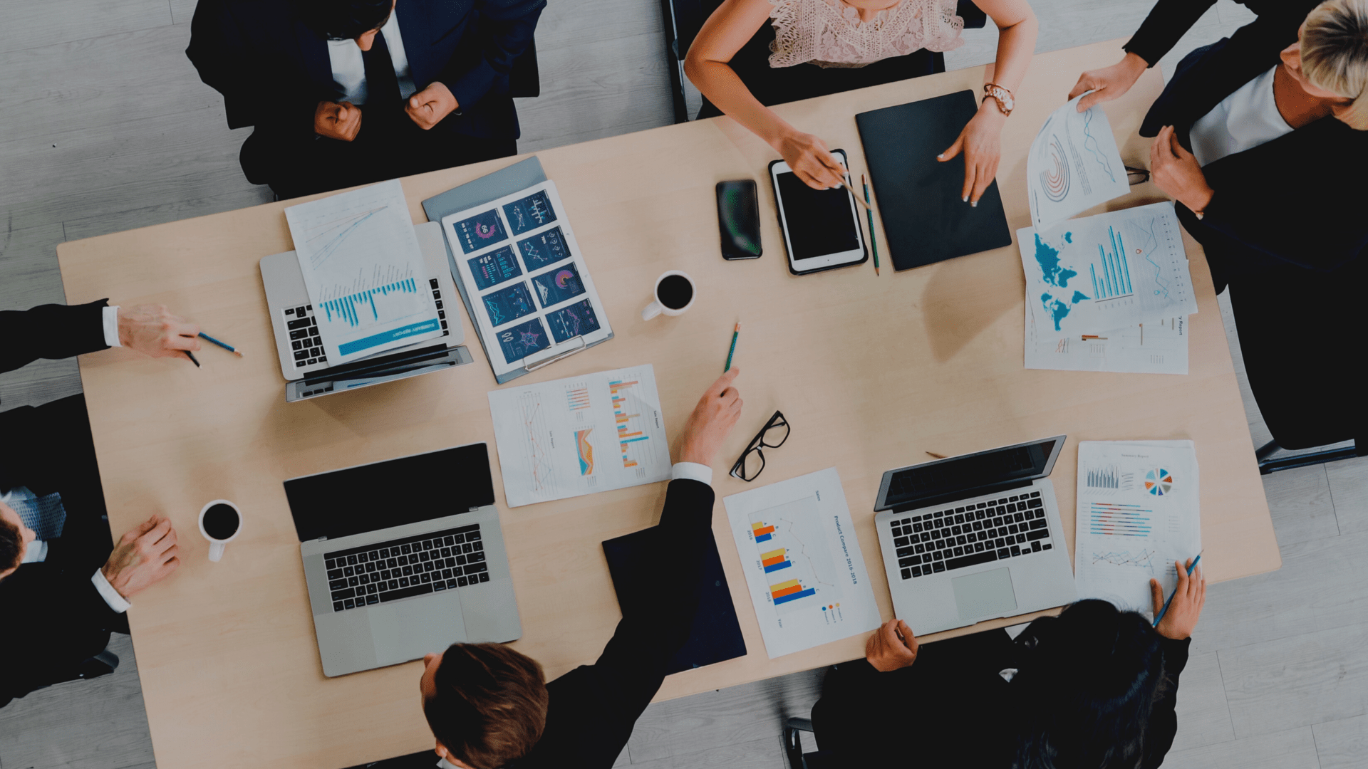 Business meeting with six people around a table, viewing laptops and documents with charts and graphs, and one has a tablet.