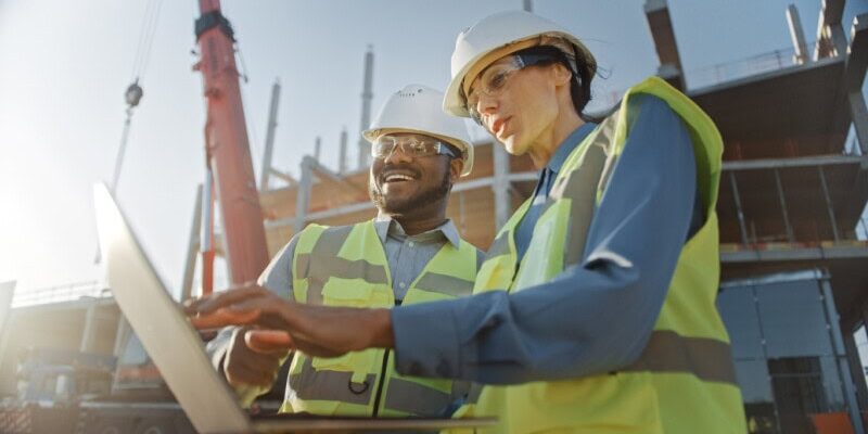 Two construction workers wearing safety gear review construction expenses on a laptop at an active construction site with a crane and building framework in the background.