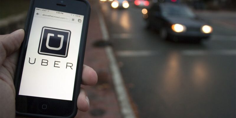 A hand holding a smartphone displaying the Uber app in front of a blurred street with moving cars at dusk.