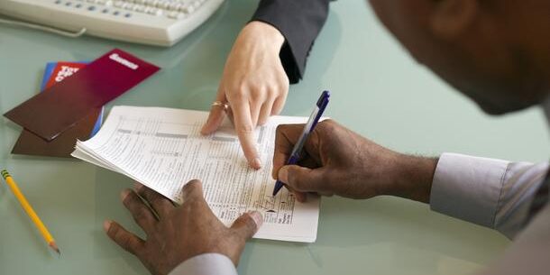 Two people reviewing and signing documents at a desk, with a keyboard, envelopes, and a pencil nearby.