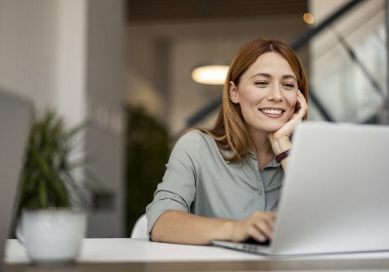 A woman with red hair sits at a desk, smiling while looking at a laptop. looking up Accounting software integrations that integrate seamlessly with ERP software