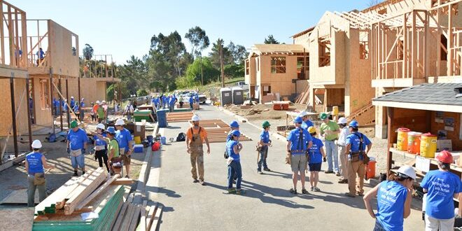 A group of people wearing blue shirts and hard hats work on multiple house constructions under a sunny sky.