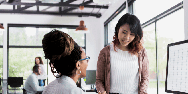 Two women discuss work at a desk in a modern office with large windows, while other colleagues work in the background.