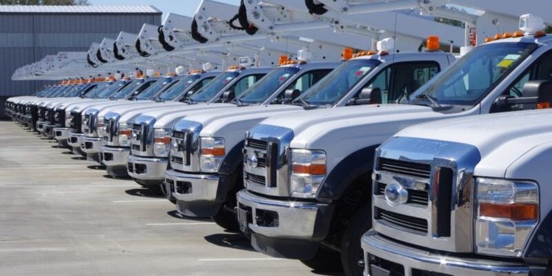 A row of white utility bucket trucks parked in a lot, with hydraulic lifts extended.