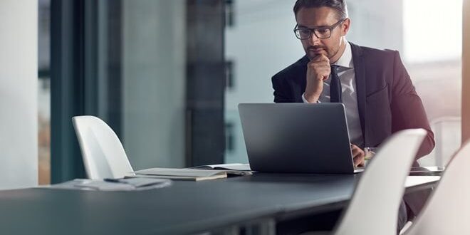 A man in a suit and glasses sits at a conference table, looking at a laptop.