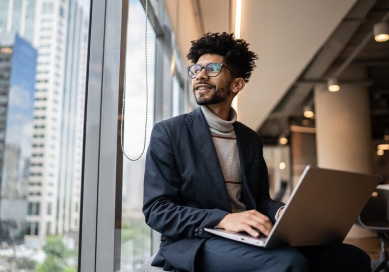 A man in business attire sits by a window in a modern office, using a laptop and choosing the right expense management application