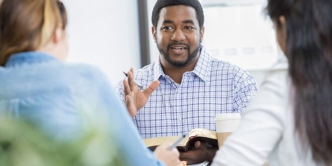 Man speaking to two people at a table, holding an open book, with a coffee cup nearby.