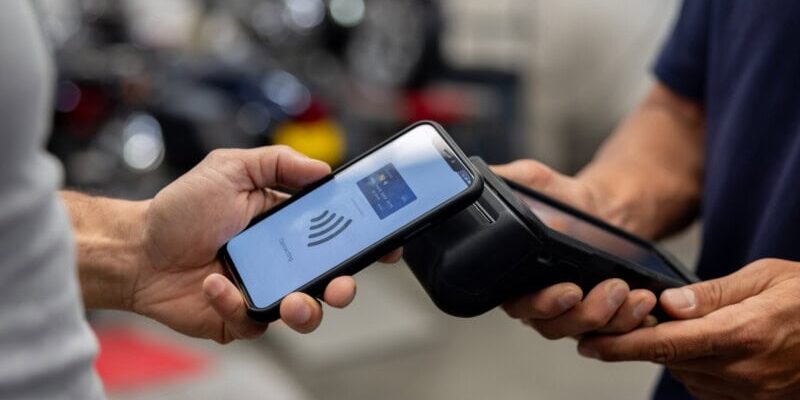 A person holds a smartphone near a card reader for contactless payment, while another person holds the terminal.