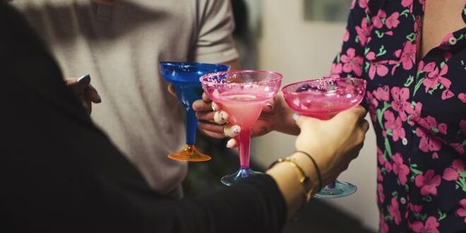 Three people holding colorful margarita glasses filled with pink drinks, toasting at a casual gathering.