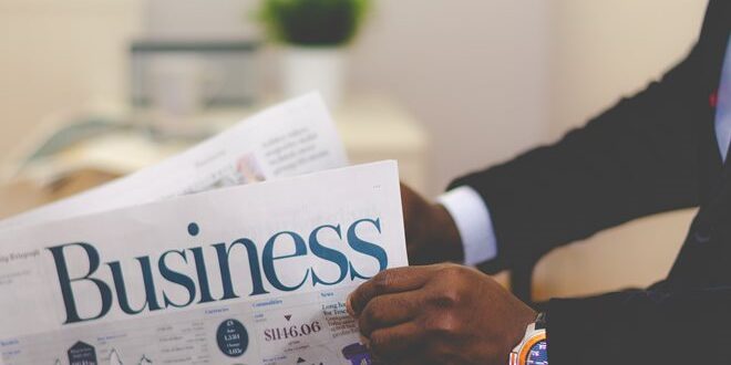 Person reading a business newspaper, wearing a dark suit and a wristwatch, with a plant in the background.