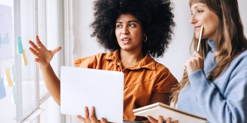 Two women stand by a window, collaborating; one gestures while holding a laptop displaying financial controls, the other holds a notebook and pen, both facing sticky notes on the glass.