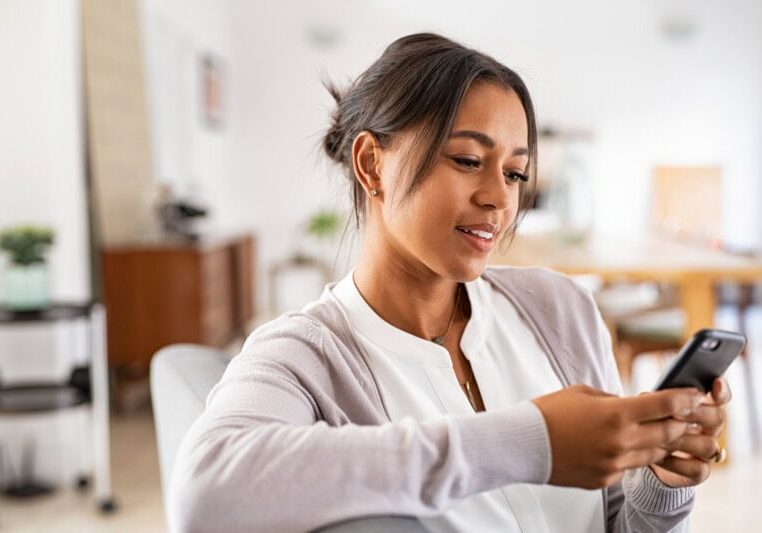 Woman sitting indoors, smiling slightly while looking at her smartphone, wearing a white blouse and light gray cardigan, perhaps reviewing nonprofit budgeting tips on her device.