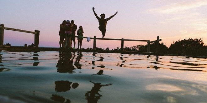 Person jumping into water near a group of people standing by a railing at sunset.