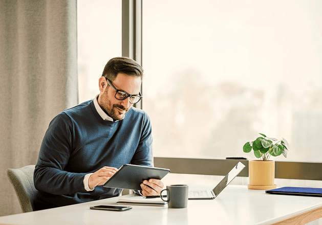 A man in glasses sits at a desk by a window using a tablet with spend management software. A laptop, smartphone, mug, and small potted plant are arranged neatly on the desk.