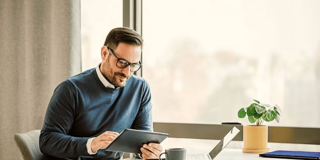 A man in glasses sits at a desk by a window using a tablet with spend management software. A laptop, smartphone, mug, and small potted plant are arranged neatly on the desk.