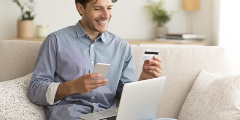 A man sits on a sofa holding a smartphone and a credit card, pondering corporate credit card limits as he faces an open laptop.