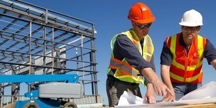 Two construction workers in safety gear review blueprints at a building site with a metal structure in the background.